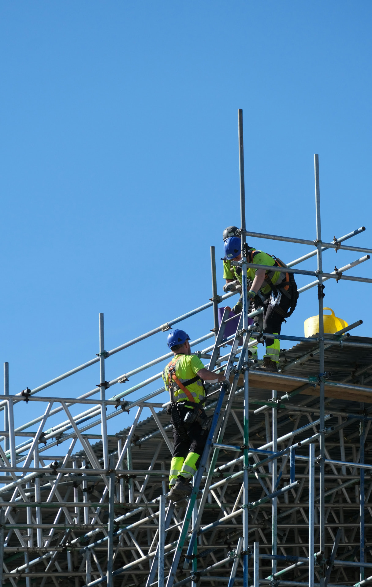 Two scaffolders in hi-vis and blue hard hats working on an aluminium scaffold structure against a clear blue sky