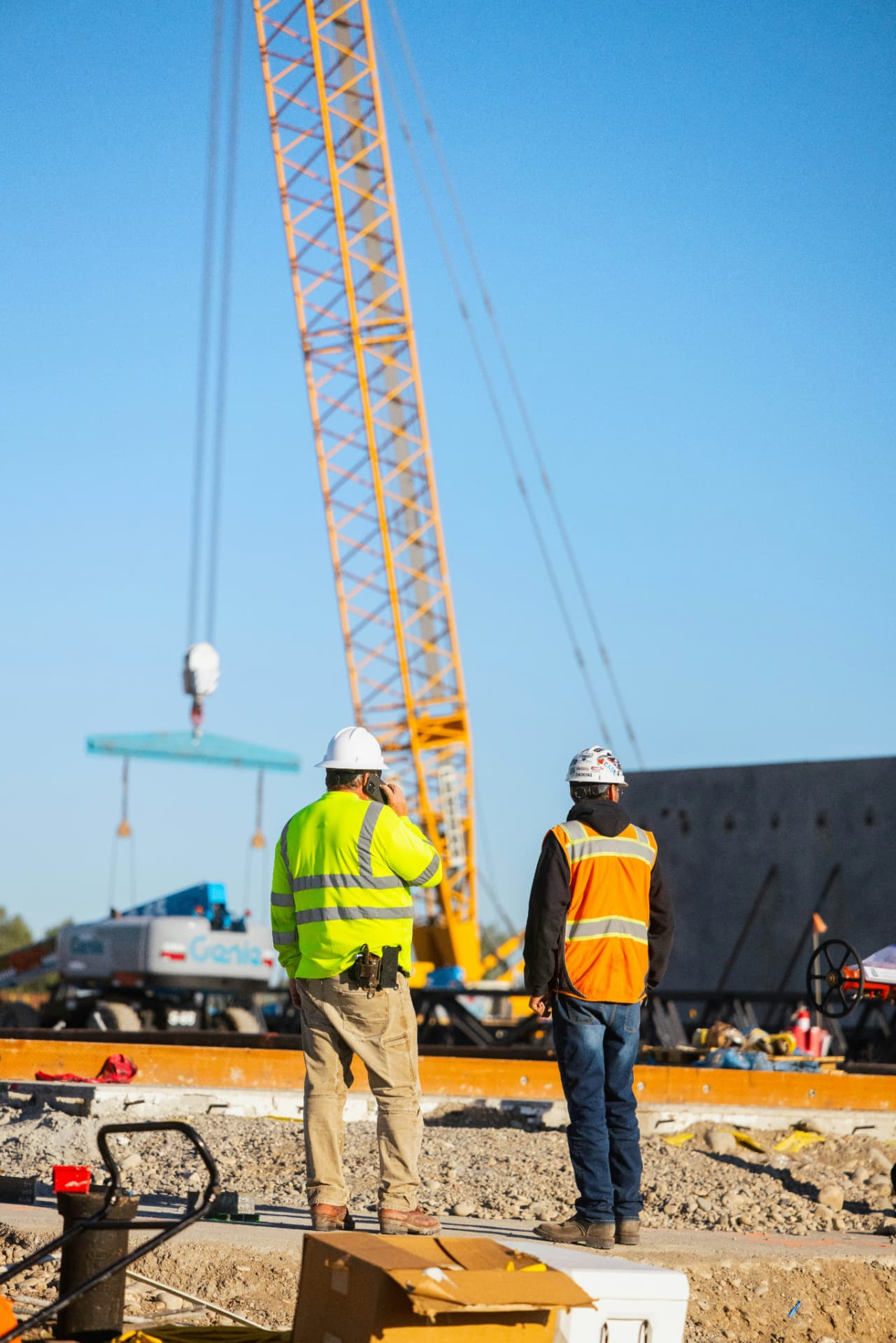 Two construction workers on site, one on the phone, watching a crane lift
